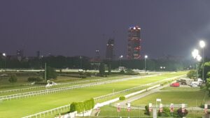 SAN SIRO_PISTA_VISTA AEREA IN NOTTURNA | AGIMEG Night view of a lit horse track with green fields and white rail fences, and a distant city skyline with tall, illuminated buildings under construction.