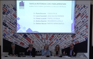 Scommettere il Futuro Forum Famiglie | AGIMEG Panel of six people at a roundtable press conference; a projection screen behind reads 'TAVOLA ROTONDA CON I PARLAMENTARI' in Italian.