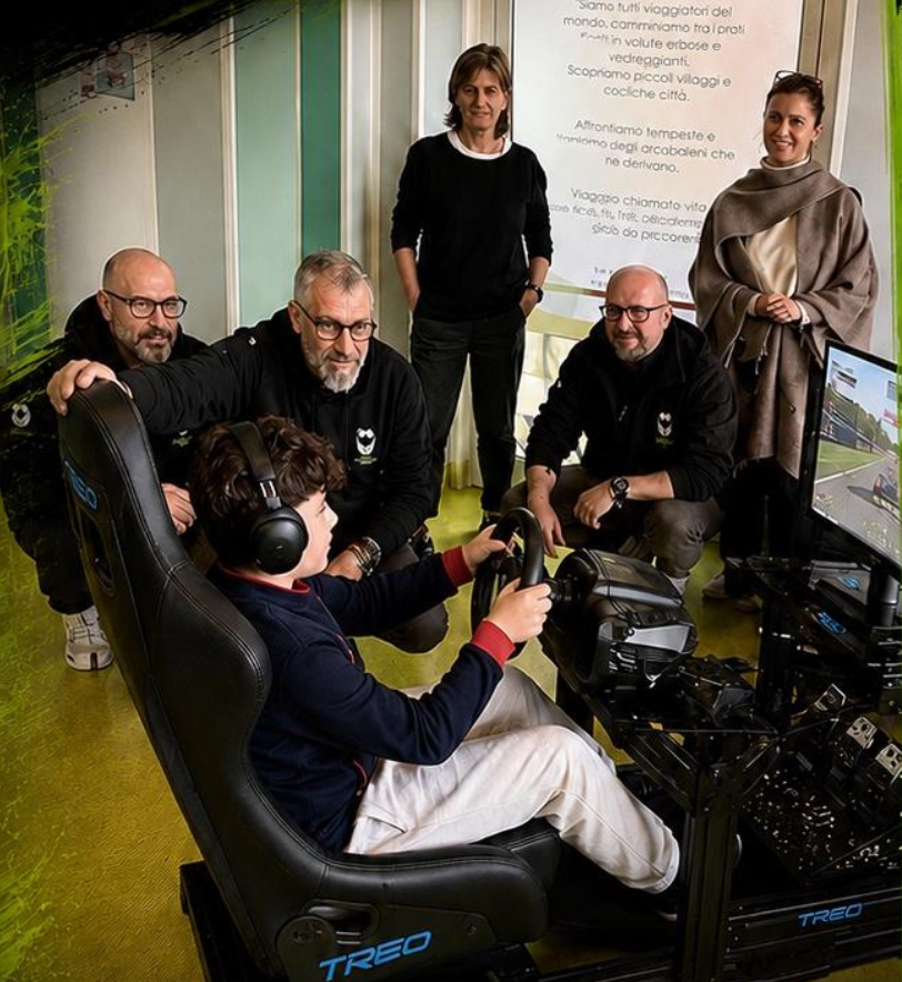 Staryes ospedali | AGIMEG Young person wearing a headset sits in a racing simulator seat, hands on a steering wheel, while four adults in black hoodies stand and watch nearby.
