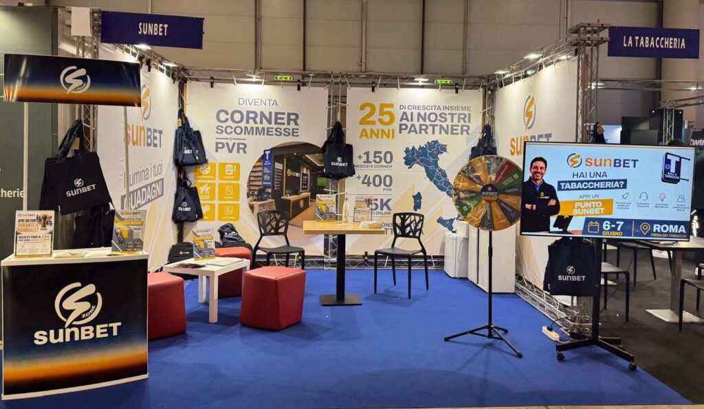 Sunbet Tabaccai | AGIMEG Sunbet exhibition booth at a trade show with banners, tote bags, and a spinning prize wheel on a blue carpeted floor.