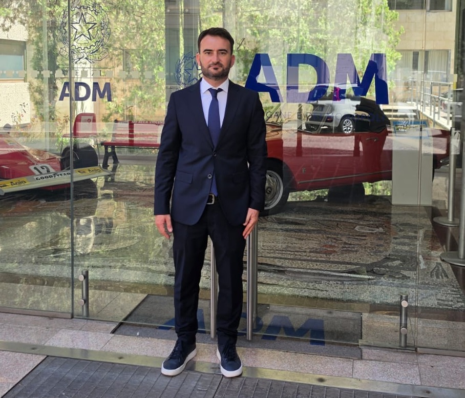 Vito Palo | AGIMEG Man in a navy suit and blue tie standing outside a glass entrance with ADM logo visible on the doors and windows, reflection in glass.